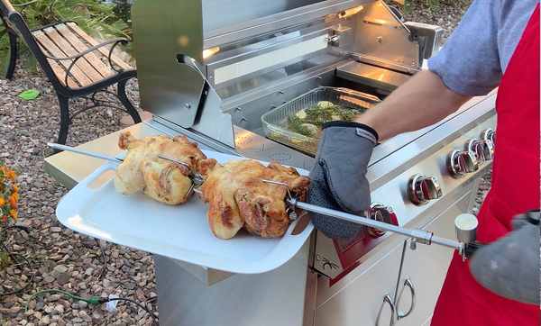 “Person wearing oven mitts holding a platter beneath two rotisserie chickens beside a KoKoMo grill.”