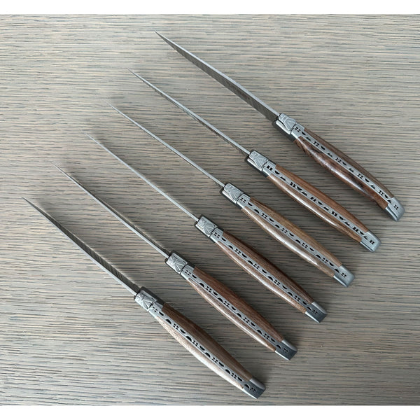 Six walnut-handled knives arranged diagonally on gray wood, showing decorative metal spines and bee emblems