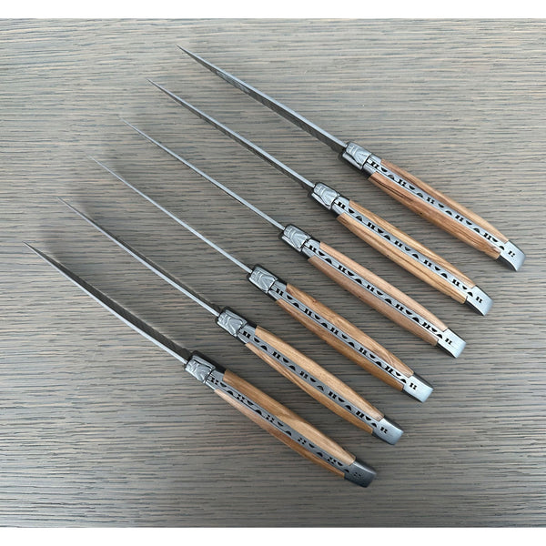 Six olive wood-handled knives arranged diagonally on gray wood, showing decorative metal spines and bee emblems