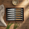 Olive wood steak knife set in a brown presentation box on a rustic table beside a linen napkin, herbs and a ceramic bowl