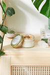 Jar of candle with lid open on a wooden surface with plants in the background