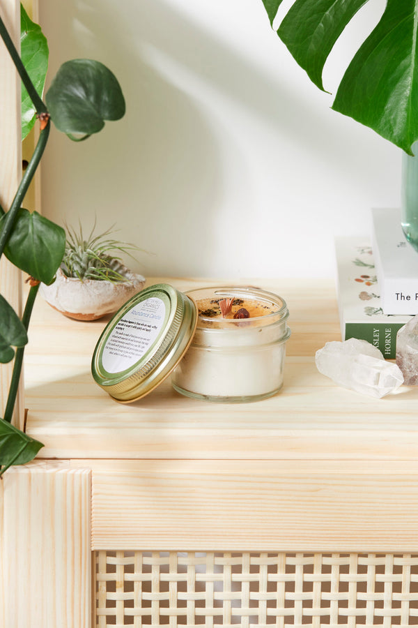Jar of candle with lid open on a wooden surface with plants in the background