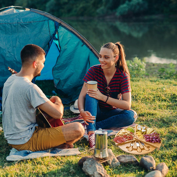 Two people camping by a lake, one playing guitar and the other holding a coffee cup, with a picnic spread out on the grass.