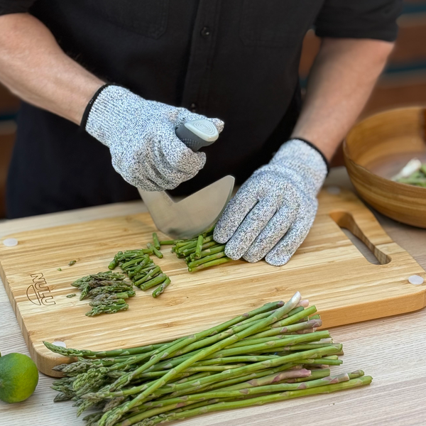 Person wearing gray NULU cut-resistant gloves while chopping asparagus with a curved knife on a wooden cutting board