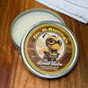 Open tin of Dr. B.Strong’s Board Balm on a wooden surface, with pale balm and bee-illustrated lid beside a white cloth
