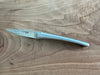 Laguiole en Aubrac matte steel steak knife resting diagonally on wood, showing the blade and tapered handle