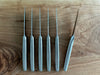 Six matte stainless steel steak knives arranged side by side on wood, showing their slim blade profiles