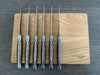 Six ash-handled steak knives arranged on a wooden box lid, showing their decorative metal spines and slim blade profiles