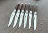 Six Laguiole en Aubrac steak knives arranged side by side with branded steel blades and ash wood handles