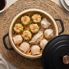 Overhead view of an Alva bamboo steamer filled with assorted dumplings, with the black lid resting alongside