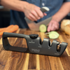 Black NULU adjustable knife sharpener on a wooden cutting board, with a person slicing cucumber in the background