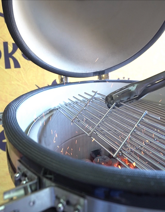 Tongs lifting the hinged cooking grate above glowing charcoal inside an open KoKoMo Kamado grill