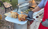 “Person wearing oven mitts holding a platter beneath two rotisserie chickens beside a KoKoMo grill.”