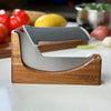 NULU acacia knife dock holding a curved NULU knife on a kitchen counter with vegetables in the background