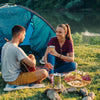 Two people camping by a lake, one playing guitar and the other holding a coffee cup, with a picnic spread out on the grass.