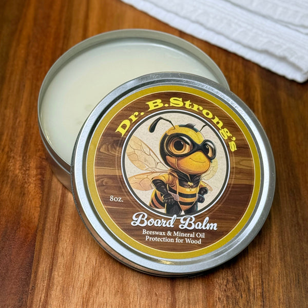 Open tin of Dr. B.Strong’s Board Balm on a wooden surface, with pale balm and bee-illustrated lid beside a white cloth