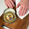 Person buffing a wooden cutting board with a white cloth beside an open tin of Dr. B.Strong’s Board Balm