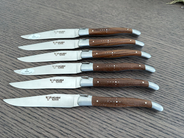 Six Laguiole en Aubrac ash wood steak knives arranged diagonally on a gray wooden surface