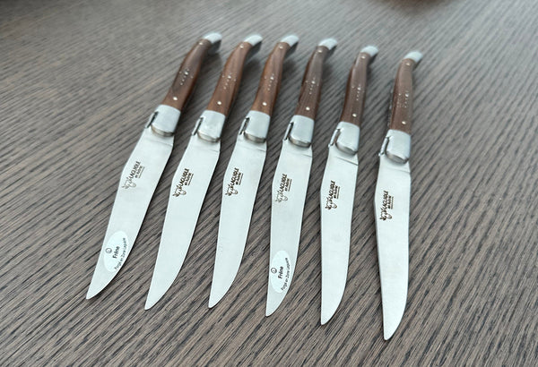 Six Laguiole en Aubrac steak knives arranged side by side with branded steel blades and ash wood handles