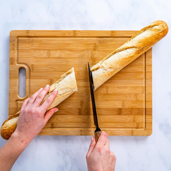 Person slicing a baguette on a wooden cutting board with a knife.