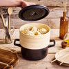Hand lifting the lid from an Alva Dutch oven and stacked bamboo steamer filled with dumplings on a wooden table